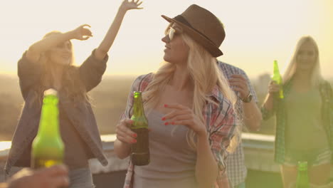 A-young-female-in-glasses-and-a-hat-moves-in-a-dance-with-her-friends-on-the-roof.-They-celebrate-a-rooftop-party-with-beer-from-colorful-green-bottels.